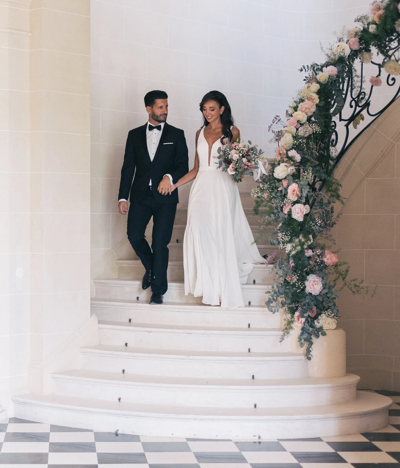 Couple de mariés descendant un escalier décoré d’une imposante composition florale pastel — L’Atelier d’Olivier