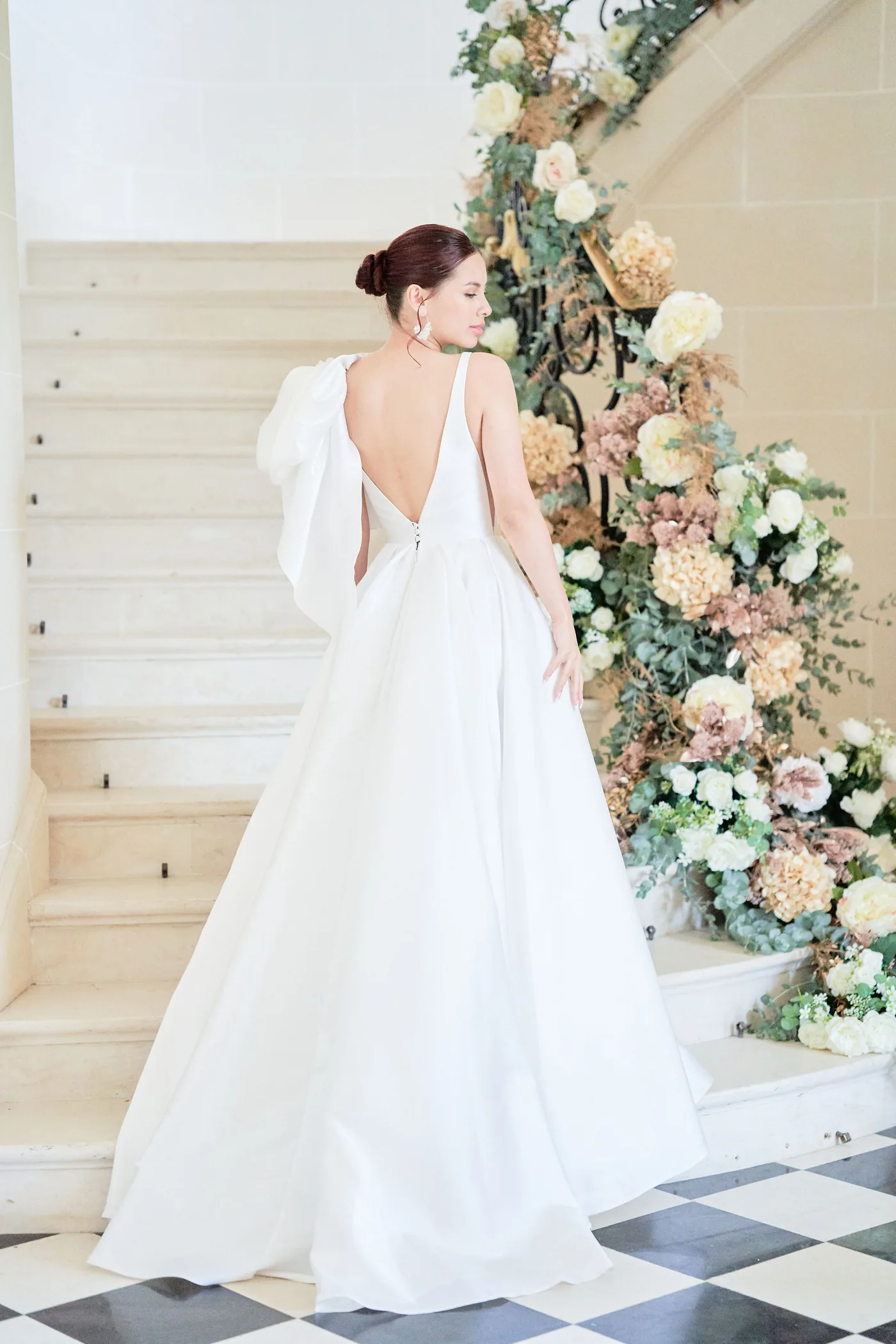 Mariée en robe blanche devant un escalier abondamment décoré de fleurs pastel — L’Atelier d’Olivier